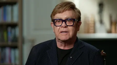 BBC Sir Elton John speaking to Laura Kuenssberg, who is behind the camera. He has ginger hair and blue eyes. He is wearing a black t-shirt, black blazer, and thick, square, black-rimmed glasses.