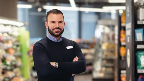 Marks & Spencer M&S Chief executive Stuart Machin in navy jumper and shirt, standing with arms folded inside store