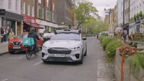 BBC A white Ford Mach-E is shown driving down a street in London. A Deliveroo courier is cycling past the left side of car in the opposite direction. The right side of the road is flanked by bushes and flower boxes, which form a barrier between chairs and tables of restaurants' outdoor dining areas on pavements. The BT Tower is visible in the very background.