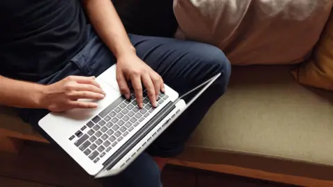 Getty Images An unrecognisable man wearing a navy t-shirt and dark blue jeans is sitting on a sofa using a laptop.