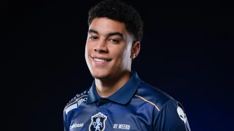 Team Liquid A man wearing a blue polo shirt with logos on it smiles at the camera against a black background