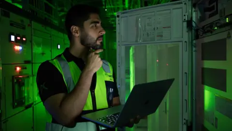 Khazna A dark-haired man with a beard stands in front of a large computer in a data centre in the UAE. He holds an open laptop in his left hand and touches his beard with his right. The background is lit by a green glow from the surrounding equipment.