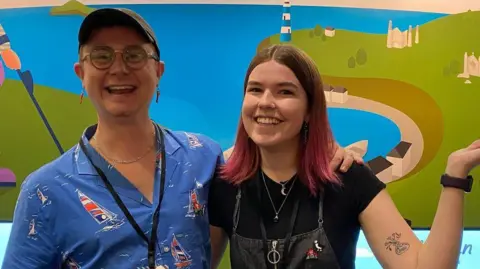 Studio Morgan A young man and young woman stand in front of a large, colourful cartoon picture of a shoreline. Both are smiling. The young man wears glasses, a shirt with a windsurfer pattern on it and a baseball cap. The woman wears dungarees over a black t-shirt. The ends of her hair are dyed pink.