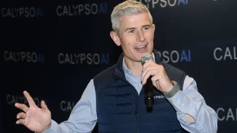 CalypsoAI Donnchadh Casey, wearing a company branded gilet speaks at a conference.