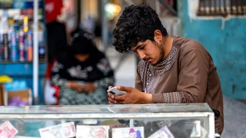WAKIL KOHSAR/AFP via Getty Images A man sitting at a money exchange stall playing on his phone.
