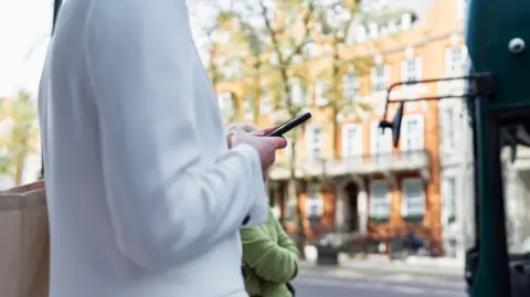 Getty Images Woman in white cardigan looks at mobile phone in a London street