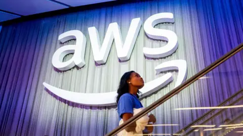 Getty Images A woman walking up stairs in front of a giant AWS sign. It is the three letters AWS with an Amazon smiley-face-like arrow underneath.