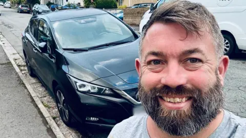 Kerry Dunstan Bearded Kerry Dunstan stands in front of his black Nissan Leaf
