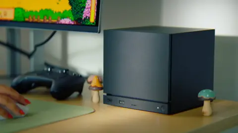 Valve A large black cube sits on a desk. It has a strip cut out near the bottom, below which is a power button and three USB ports.