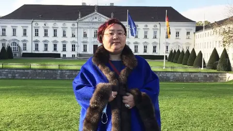 Metropolitan Police Qian Zhimin - wearing glasses and her hair dyed red - standing in front of a building in Berlin. She is wearing a fur-lined bright blue coat. 