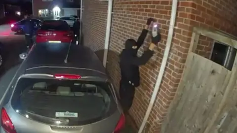 Abbie Brookes-Morris A CCTV image showing a person dressed in black clothing and wearing a balaclava holding a device against a brick wall. The individual is standing in a residential driveway next to a silver car parked close to the wall, with other vehicles visible in the background under street lighting. A wooden fence is partially visible on the right side.