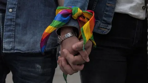 Getty Images A same-sex couple, dressed in denim jackets, hold hands during an event to raise awareness of gay rights. Their wrists are tied together by a rainbow ribbon.