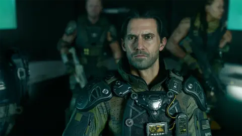 Activision A screenshot shows a realistic rendering of actor Milo Ventimiglia's character wearing an elaborate military uniform and body armour. He's looking up, his face illuminated by the glow of a screen, as his squadmates look on in the background.