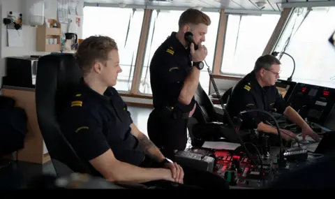 Three men in the Swedish coastguards uniform sit at the control desk of a ship. Two on the outside are sitting down while the man in the middle is standing and speaking on the phone.