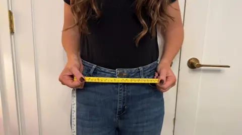 BBC A woman in jeans stands with a yellow tape measure across her waist.