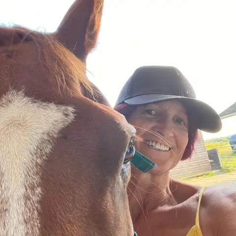 BBC Sue - a woman smiling while wearing a baseball cap - stands next to a horse. The horse is closer to the camera, with only it's eye and forehead visible beside her.