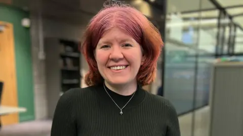 Sarah Blackburn has shoulder-length pink and orange hair. She is smiling at the camera and wearing a black top and a silver necklace