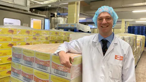 Stuart Louden wears a white protective coat and a food-hygiene hat. He's leaning on a pallet loaded with boxes of Tunnock's wafer bars.