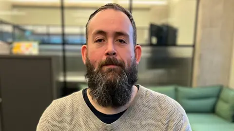 Jamie Trimmer has short, dark hair and a long, dark beard with grey through it. He is wearing a grey jumper. He is sitting in what looks like an office space