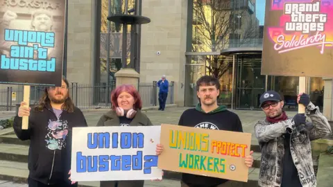 BBC Four people, three men and a woman, hold signs outside a building. Their signs read "unions ain't busted" and "union busted?" and "unions protect workers" and "grand theft wages solidarity"