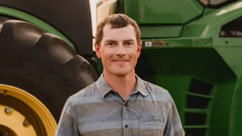 Jake Leguee Smiling and wearing a stripey blue and grey shirt, Jake Leguee stands in front of large tractor.