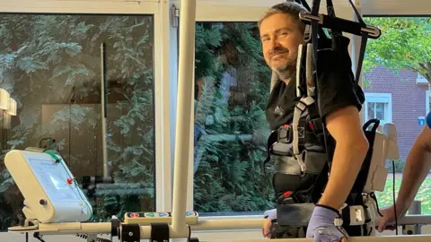 Anna Thomas Dan Richards using a machine to walk. He is standing and smiling, as he wears a harness and belts around his body.