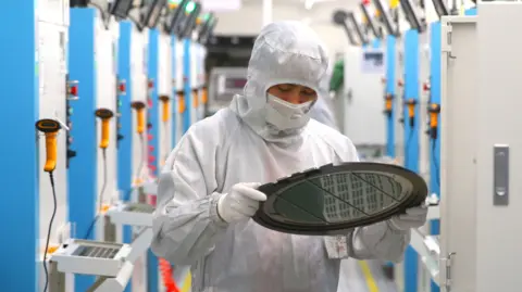 Getty Images A worker in a white protective suit inspects a silicon wafer - a round disc.