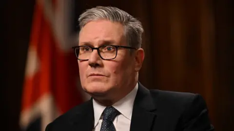 BBC Sir Keir Starmer with a serious expression pictured from the shoulder up, wearing a tie and suit, with neat black rimmed glasses, with a union flag out of focus behind him 