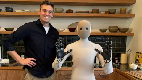 BBC NEO the humanoid robot from 1X and reporter Joe Tidy pose for a photo. Neo is a 5ft robot with soft coverings over it's robot body.