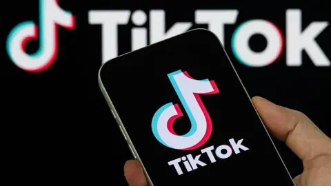 Getty Images A smartphone held in someone's hand displays the TikTok logo, with a backdrop of the company's logo behind it