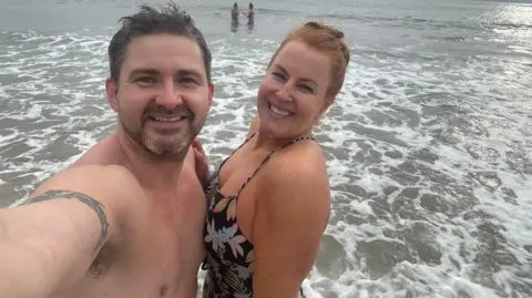 Anna Thomas A selfie of Dan (left) and his partner Anna (right) standing in the sea. Dan has short, dark hair and and Anna's red hair is tied back. They are both smiling. There are two people in the background also in the sea.