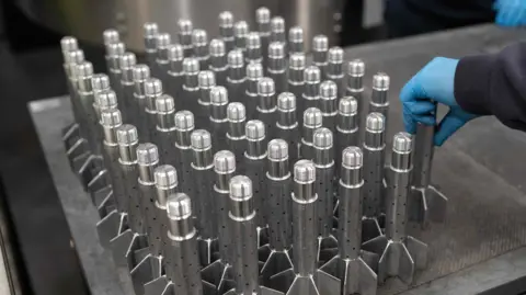 Getty Images The tail sections of around 60 mortar munitions are lined up on a table. They look like small rockets. 