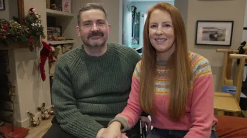 Dan and Anna smiling in their living room. Dan (left) is wearing a green jumper and Anna (right) is in a pink winter jumper with yellow and blue pattern.