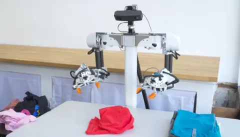 A robot with orange pincers for hands stands over a pile of laundry. The bot has a shoebox-like head with cameras and a basic frame body.