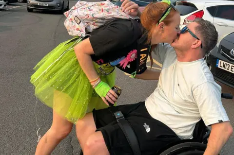 Anna Thomas A man with brown hair wearing a white t shirt, black shorts and sunglasses, sat in a wheelchair in a car park with cars in the background. A woman with red hair, dressed in a black t shirt and neon green tutu is leaning down and kissing him.