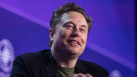 Getty Images Elon Musk stares towards the camera with a smile while sitting against a purple background on a stage