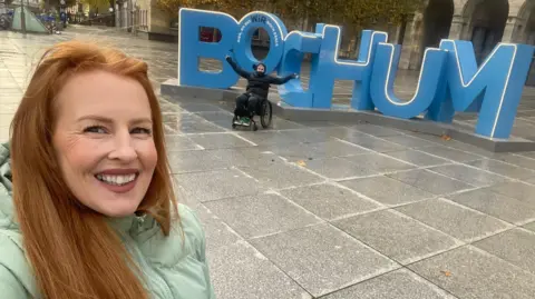 Anna Thomas A selfie of a woman with long red hair, wearing a teal puffer jacket and smiling at the camera. Behind her are large blue letters spelling BOCHUM, and in front of them is a man, dressed in all black and sat in a wheelchair with his arms up in the air.